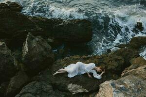 lindo jovem mulher dentro grandes branco vestir molhado cabelo deitado em uma rochoso penhasco panorama foto