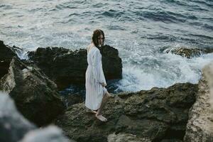 bonita mulher dentro branco vestir carrinhos em a pedras oceano natureza liberdade foto