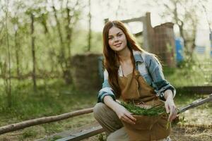 uma jovem mulher feeds dela galinhas em a Fazenda com grama, vestindo uma simples xadrez camisa, calça e avental, e sorridente para a Câmera, carinhoso para a animais foto