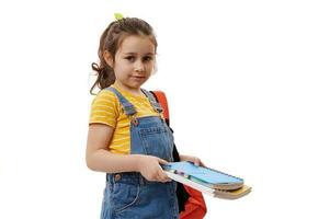 lindo criança garota, primeiro motoniveladora carregando mochila e estudando livros, olhando às Câmera, isolado em branco fundo foto