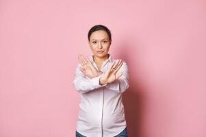 confiante grávida mulher mostra Pare sinal, cruzando braços, olhando às Câmera, expressando negação recusa em Rosa fundo foto