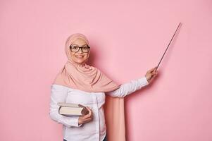 grávida muçulmano mulher professor dentro Rosa hijab e Óculos, sorridente às Câmera, apontando às cópia de espaço, explicando lição foto