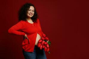 lindo multi étnico grávida mulher sorridente às Câmera, posando com uma ramalhete do vermelho tulipas para mãe ou mulheres dia foto