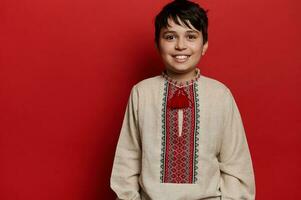 retrato do uma sorridente caucasiano adolescente Garoto dentro ucraniano tradicional bordado linho camisa, isolado em vermelho fundo foto