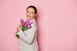 positivo mulher segurando ramalhete do roxa tulipas, sorridente olhando através dela ombros em cópia de espaço, em Rosa fundo foto