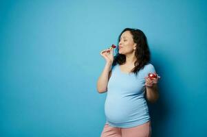 feliz grávida mulher comendo morangos em azul fundo. equilibrado dieta durante gravidez. maternidade saúde conceito foto