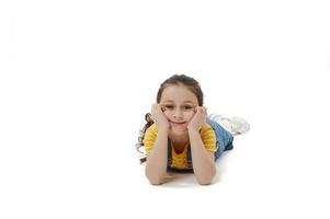 caucasiano pequeno menina dentro amarelo camiseta e jeans vestido de verão deitado em barriga, olhando às Câmera, isolado branco fundo foto