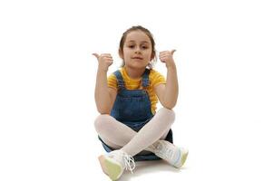 adorável criança menina dentro casual jeans roupas, olhando às Câmera, gesticulando com polegares acima, sentado em isolado branco pano de fundo foto