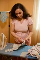 lindo grávida mulher passar roupa e dobrando recém-nascido macacões em passar roupa quadro, preparando para a nascimento do uma criança foto
