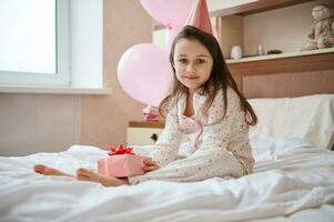 adorável aniversário menina dentro Rosa festivo chapéu, segurando presente caixa, sentado em a cama dentro dela quarto com hélio balão foto