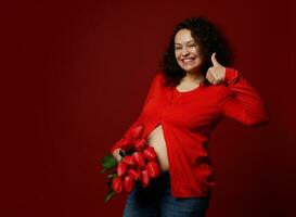 bonita grávida mulher detém uma grupo do vermelho tulipas, sorrisos, mostra polegar acima , isolado vermelho fundo. feliz mãe dia foto