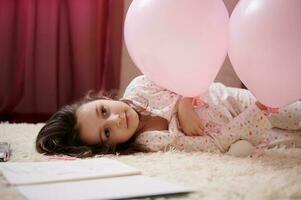 adorável delicioso pequeno criança menina dentro pijamas, deitado em uma tapete dentro dela quarto, jogando com Rosa pastel balões foto