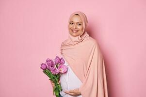 muçulmano mulher dentro Rosa hijab, segurando uma ramalhete do tulipas, tocante dela grávida barriga, sentindo-me feliz de primeiro bebê chutes foto