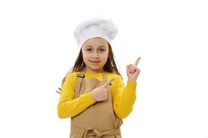 sorridente feliz criança menina vestindo chef chapéu e bege cozinha avental, pontos dedos às cópia de de Anúncios espaço em branco fundo foto