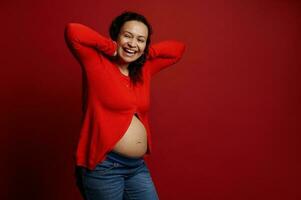 encaracolado étnico grávida adulto mulher rindo, posando nu barriga sobre isolado vermelho fundo. maternidade. pessoas. estilo de vida foto