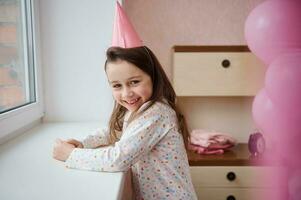 encantador pequeno menina dentro uma festivo Rosa chapéu e pijamas, carrinhos de a janela e sorrisos docemente olhando para dentro a Câmera foto