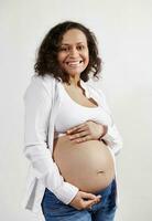 africano americano grávida mulher sorrisos às Câmera, expressando positivo emoção sentindo-me primeiro bebê chutes, fazendo carinho barriga foto