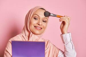fechar-se lindo meio Oriental muçulmano mulher dentro Rosa hijab, segurando sombra paleta e aplicando Maquiagem em dela face foto