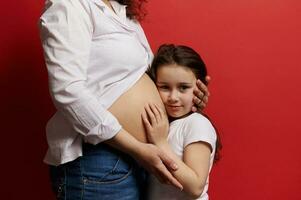 lindo pequeno menina abraçando a barriga do dela grávida mãe, olhando às Câmera, isolado em vermelho fundo. foto