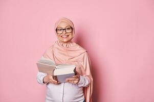 confiante lindo muçulmano grávida mulher dentro Rosa hijab e na moda copos, lendo uma livro, sorridente olhando às Câmera foto