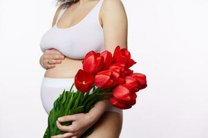 irreconhecível grávida mulher suavemente fazendo carinho dela barriga, posando com uma grupo do vermelho tulipas em branco isolado fundo foto