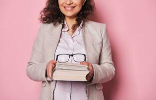 detalhes em à moda Óculos em uma livro dentro mãos do uma sorridente bonita mulher, escola professor, isolado em Rosa fundo foto