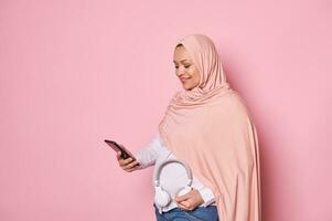 bonita muçulmano mulher dentro Rosa hijab, rolagem jogar Lista em Smartphone e colocando fones de ouvido em dela barriga foto