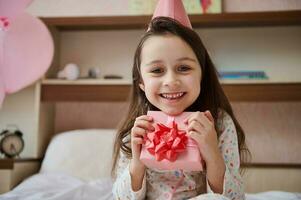 adorável pequeno criança menina dentro pijamas e Rosa festivo chapéu, abraços dela aniversário presente e sorridente olhando às Câmera foto