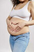 fechar-se grávida mulher colocando mãos em nu barriga, fazer coração forma a partir de dedos. feliz despreocupado gravidez 30 semana foto