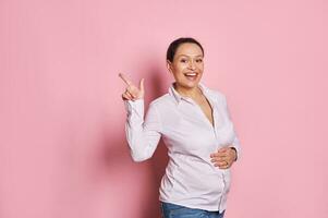 feliz grávida mulher tocante suavemente dela barriga, sorridente às Câmera, pontos dela dedo às cópia de de Anúncios espaço em Rosa fundo foto