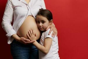 fechar-se grávida mãe suavemente abraçando dela filha quem é tocante nu barriga, isolado em vermelho fundo foto