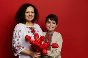 feliz sorridente étnico mulher e adolescente garoto, mãe e filho dentro tradicional ucraniano bordado camisas, segurando vermelho tulipas foto