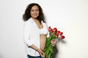 africano americano grávida bonita mulher posando nu barriga com uma ramalhete do vermelho tulipas, isolado em branco fundo foto