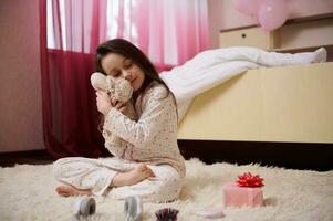 pequeno menina dentro pijamas, abraçando dela pelúcia brinquedo, sentado com dela olhos fechadas em tapete dentro dela quarto antes dia Sonhe foto