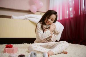 delicioso 5-6 anos velho criança menina dentro pijamas, sentado em a tapete dentro dela infantil quarto e abraçando dela pelúcia brinquedo foto