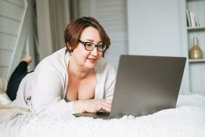 sorridente meio envelhecido mais Tamanho mulher deitado em cama, trabalhando em computador portátil a partir de casa foto