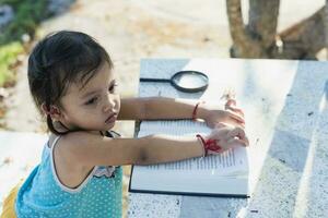 lado Visão do ásia menina sentado em uma Banco e lendo uma livro dentro a parque. foto