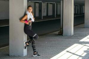 tiro do lindo fêmea corredor em pé ao ar livre segurando água garrafa e bebendo. ginástica mulher levando uma pausa depois de corrida dar certo. fique hidratado foto