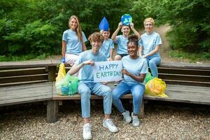 feliz voluntários segurando cartaz com 'feliz terra dia' mensagem. voluntariado, caridade, limpeza, pessoas e ecologia conceito - grupo do feliz voluntários com lixo bolsas limpeza área dentro parque foto