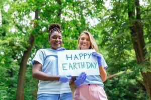 feliz voluntários segurando cartaz com 'feliz terra dia' mensagem. voluntariado, caridade, limpeza, pessoas e ecologia conceito - grupo do feliz voluntários com lixo bolsas limpeza área dentro parque foto