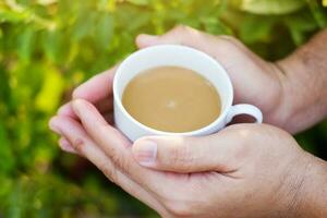 mão segurando branco cerâmico copo do quente café com natural luz manhã dentro a jardim. cafeína bebidas causa uma fresco sentimento. foto