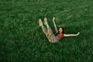 uma jovem mulher jogando jogos dentro a parque em a verde Relva espalhando dela braços e pernas dentro diferente instruções queda e sorridente dentro a verão luz solar foto