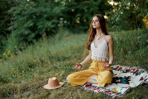 uma jovem hippie mulher medita dentro natureza dentro a parque, sentado dentro uma lótus posição em dela colorida xadrez e desfrutando harmonia com a mundo dentro roupas ecológicas foto