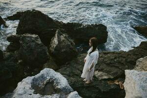 uma mulher dentro uma branco vestir com molhado cabelo carrinhos em a pedras de a oceano topo Visão foto