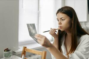 mulher segurando Maquiagem lápis dentro mão dentro de casa perto janela e olhando dentro espelho foto