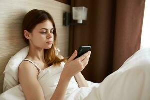 retrato do uma mulher dentro cama debaixo uma branco cobertor com uma Móvel telefone e Projeto interior foto