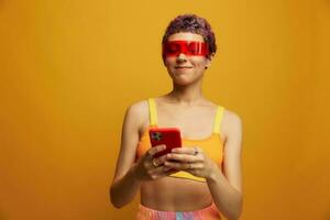 mulher blogueiro sorridente dentro incomum vermelho milenar óculos segurando dela telefone e olhando às a tela contra a laranja fundo dentro uma à moda amarelo topo foto