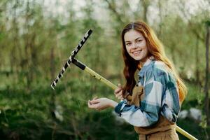 uma mulher sorridente belas e olhando às a Câmera, uma agricultor dentro trabalhos roupas e a avental trabalhando ao ar livre dentro natureza e segurando uma ancinho para reunir Relva e forragem para a animais dentro a jardim foto