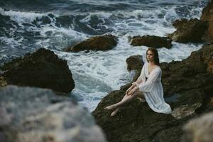 bonita mulher com molhado cabelo dentro uma branco vestir senta em uma penhasco natureza inalterado foto