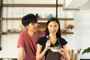 um jovem casal asiático está comendo juntos e sorrindo alegremente enquanto cozinha sua salada na cozinha.